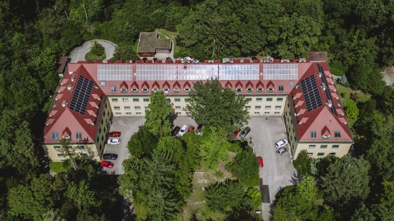 Blick von oben auf ein Gebäude mit Parkplatz und Photovoltaikanlage auf dem Dach, umgeben von Wald.