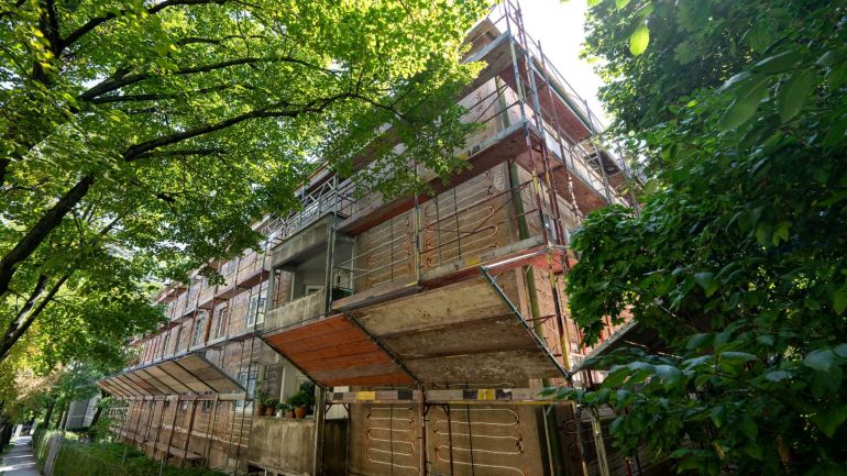 Hausfassade mit Gerüst und Leitungen, umgeben von Bäumen