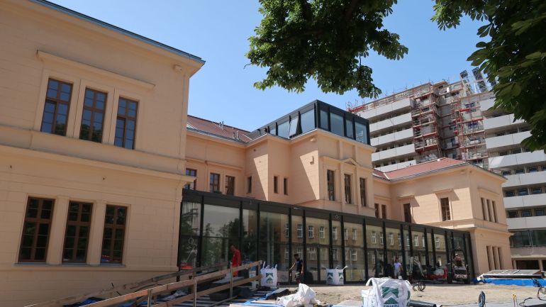 Fassade eines ehemaligen Spitalsgebäudes mit verglastem Eingangsbereich und Dachausbau.