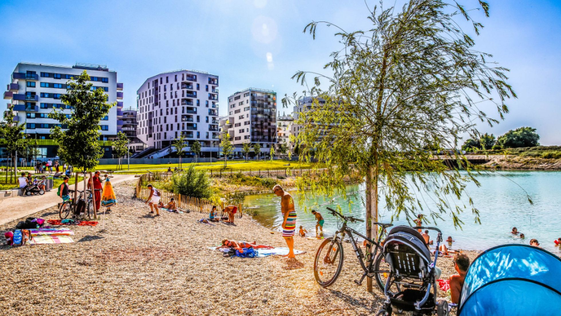 Kiesstrand am See, badende Menschen, Gebäude im Hintergrund