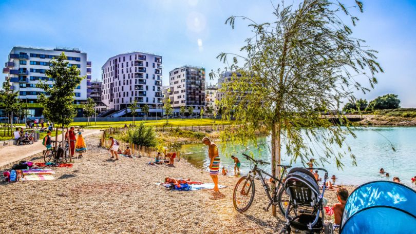 Kiesstrand am See, badende Menschen, Gebäude im Hintergrund
