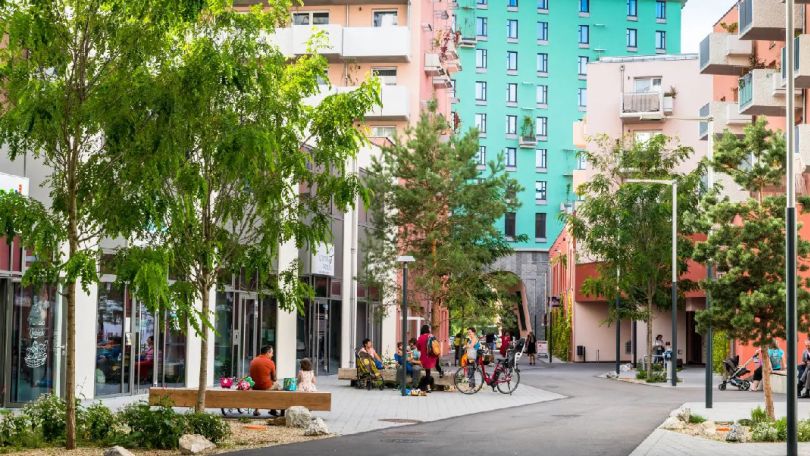 Bunte Wohnbauten, Straße, Gehwege mit Sitzmöglichkeiten, Bäume links und rechts entlang der Straße