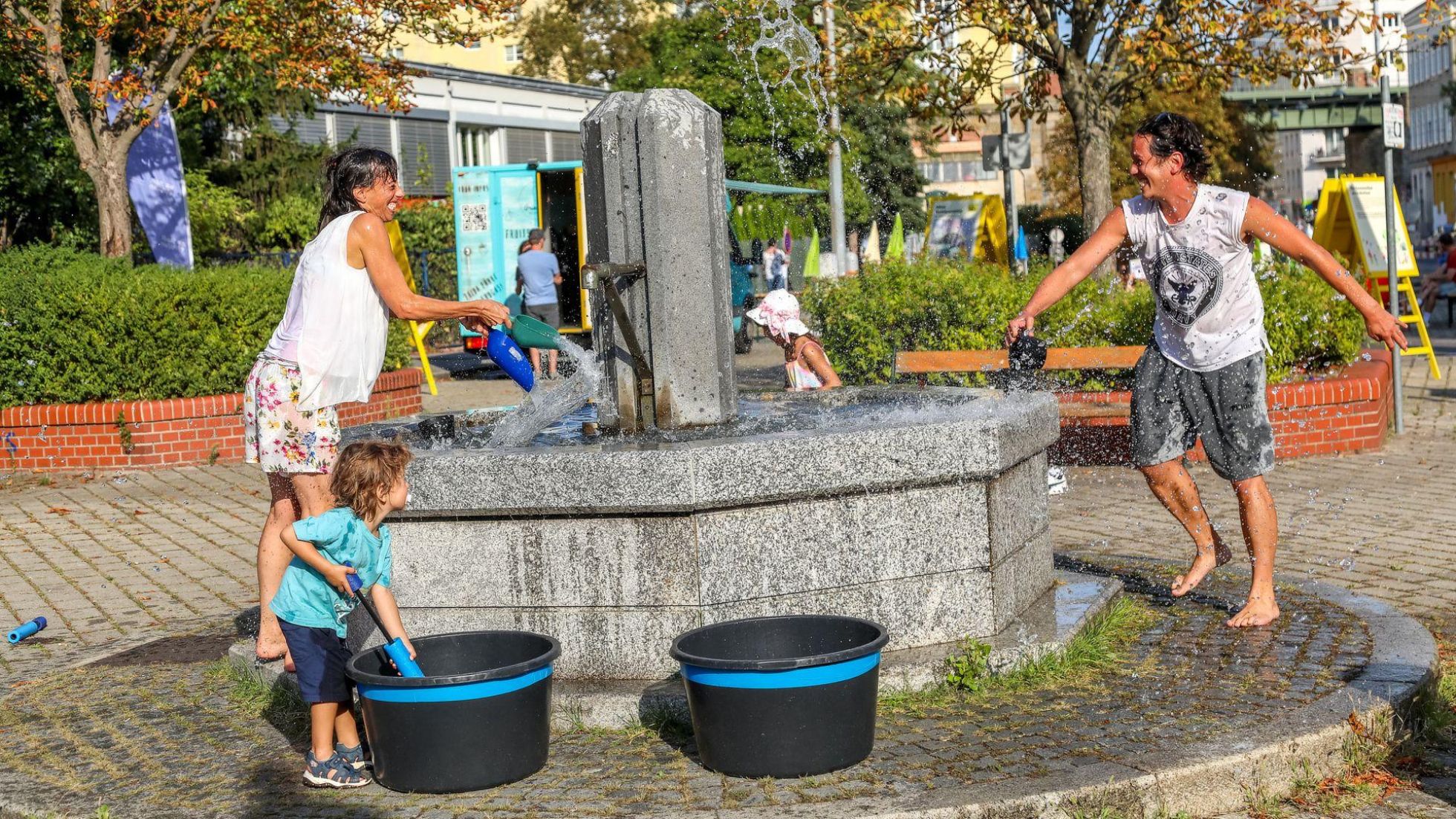 Erwachsene und Kind beim Springbrunnen, die sich gegenseitig nass spritzen.