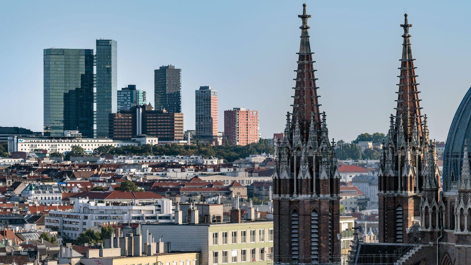 copyright: Stadt Wien / Gerd Götzenbrucker Links im Hintergrund eine Skyline mit verglasten Hochhäusern, rechts vorne 2 Kirchtürme, dazwischen Stadtgebiet.