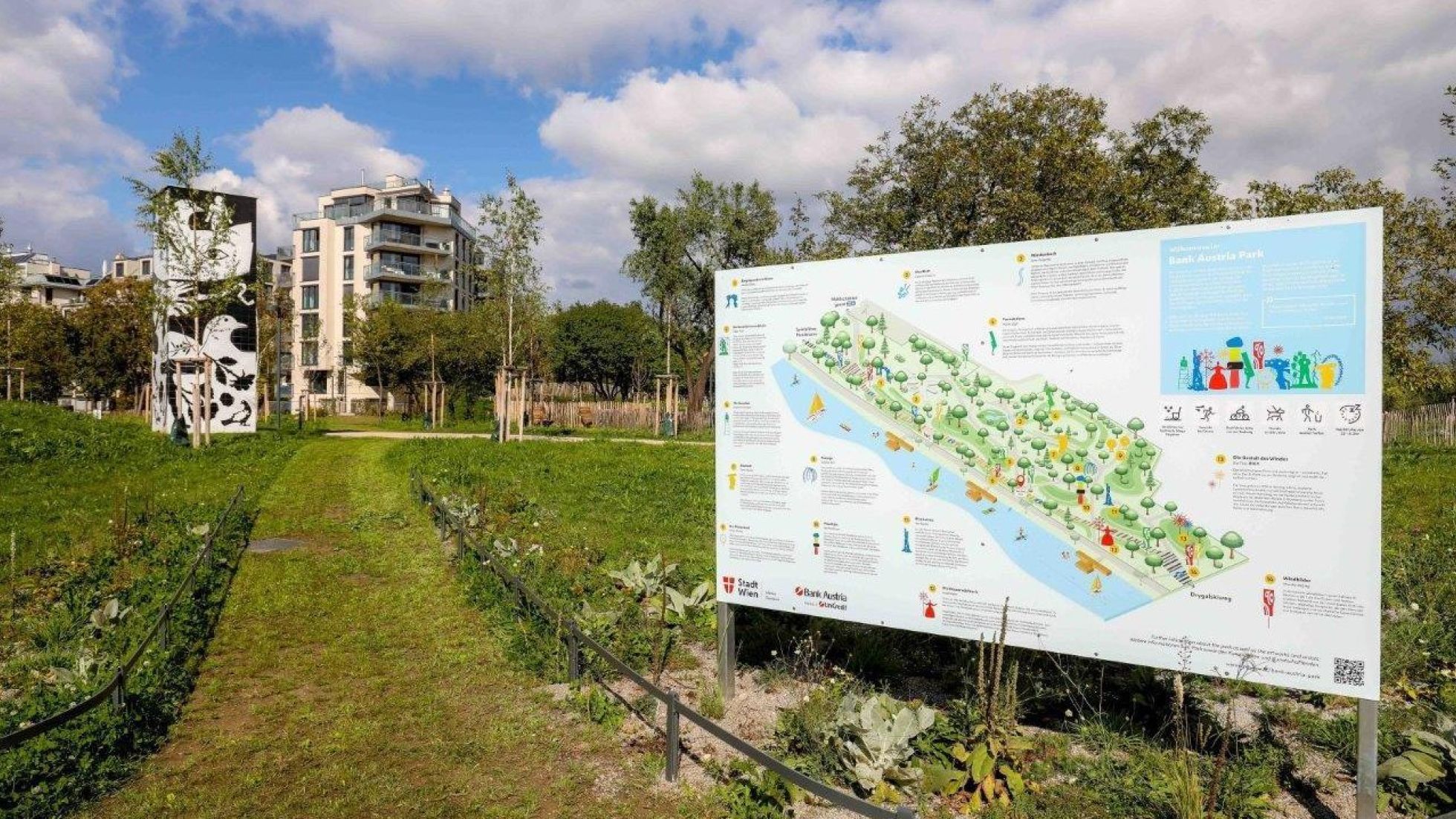 Lageplan im Park, daneben ein Weg, im Hintergrund Wohngebäude und Bäume.