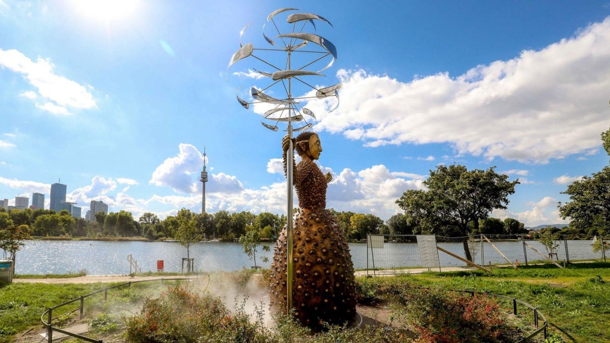 Statue mit zackigem Kleid hält Windspiel, Dampf kommt aus dem Boden, im Hintergrund die Alte Donau und die Skyline Wiens.
