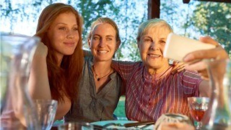 3 Frauen sitzen an einem Tisch, legen sich Arm um Schulter, die Älteste macht mit dem Handy ein Gruppen-Selfie