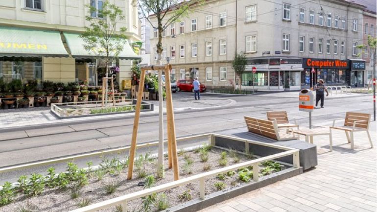 Straße mit Gleisen und Gehsteige mit Bäumen und Sitzgelegenheiten