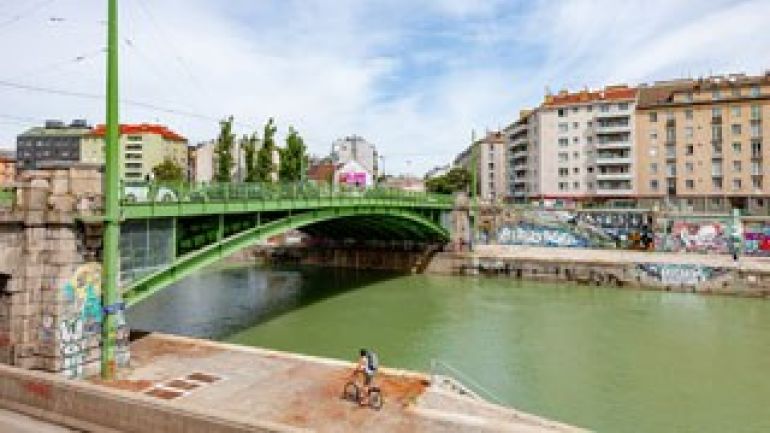 Grüne Brücke aus Metall beim Donaukanal, dahinter Wohngebäude
