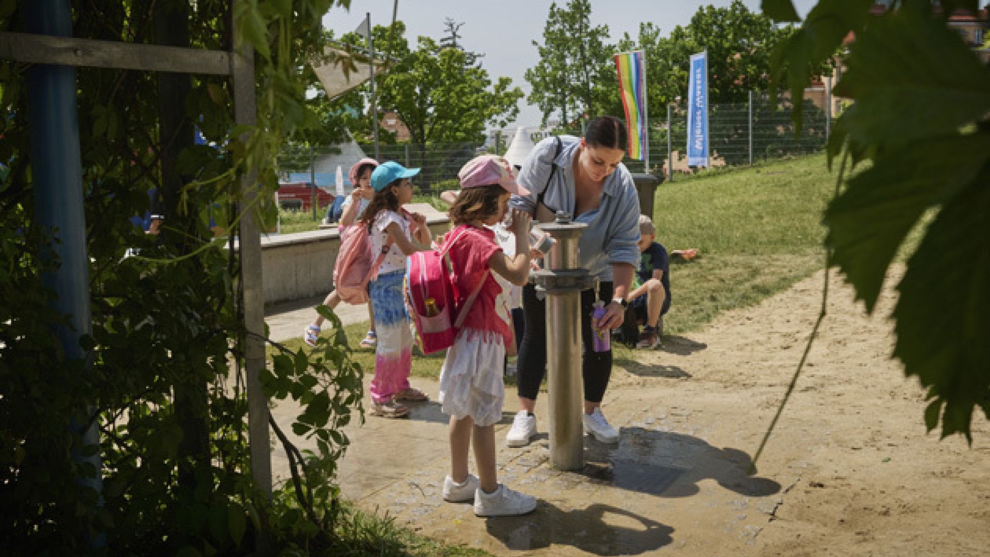 Ein Bub balanciert auf einem Surfbrett, während er auf einer roten aufblasbaren Wasserbahn gleitet. Im Hintergrund sind Zelte und Menschen zu sehen, in der Umgebung gibt es Grün und ein paar Bäume.