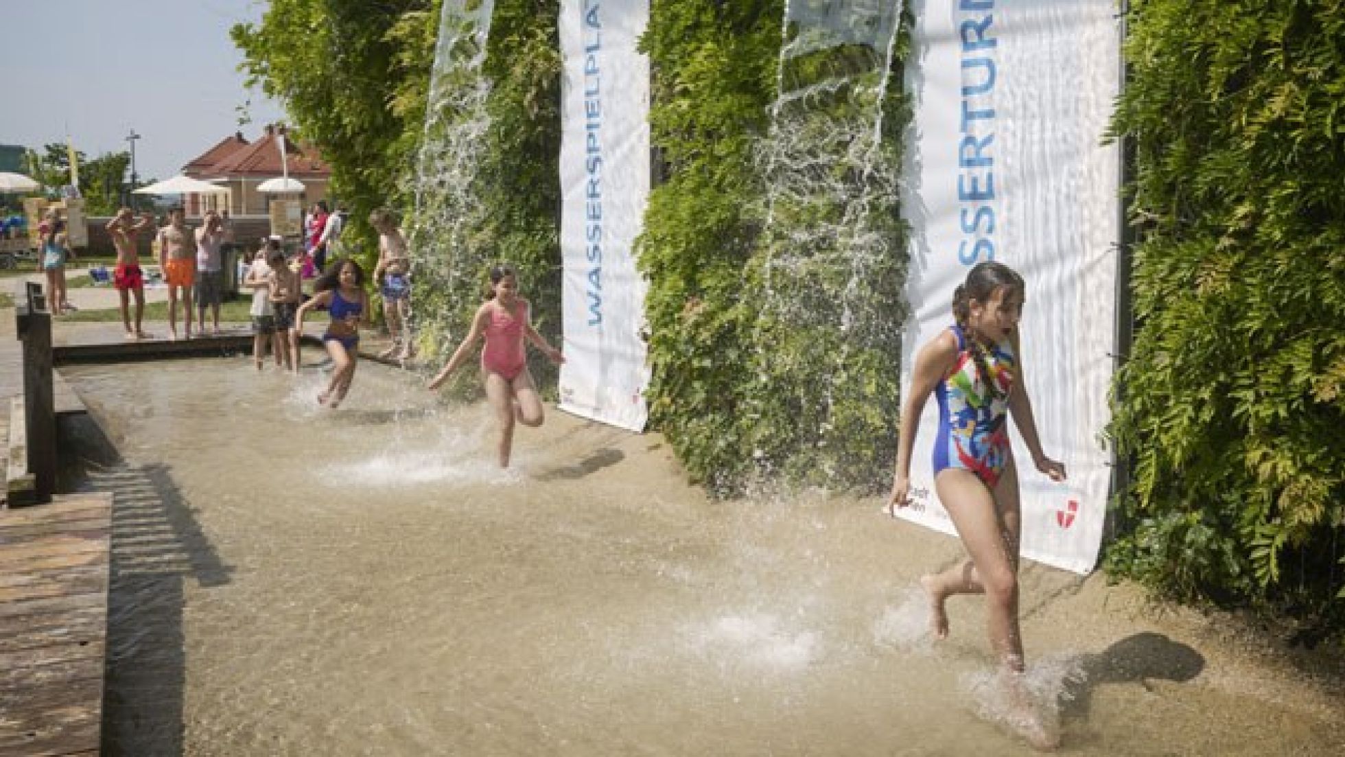 Mehrere Kinder laufen in Badebekleidung durch einen künstlichen Wasserfall.