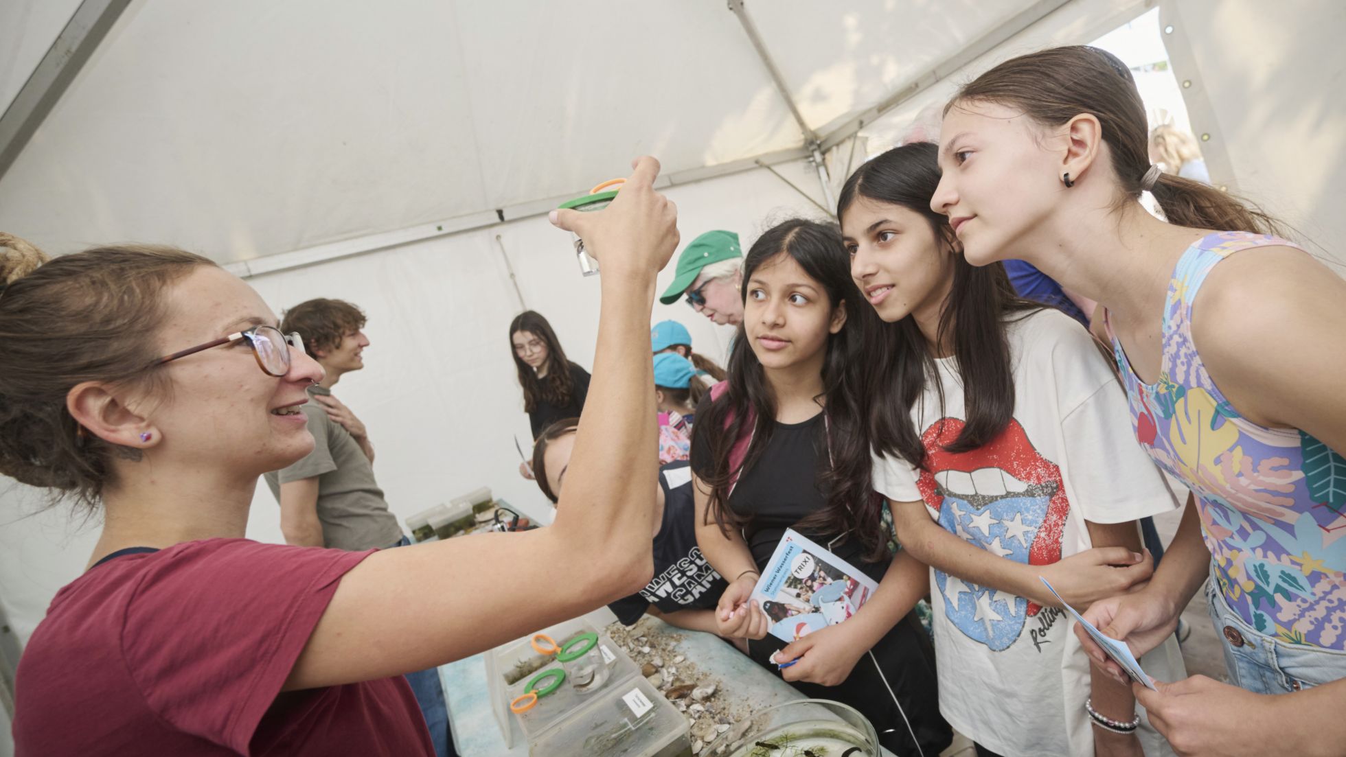 3 Mädchen beobachten einen Wasserbecher, den eine Mitarbeiterin präsentiert.