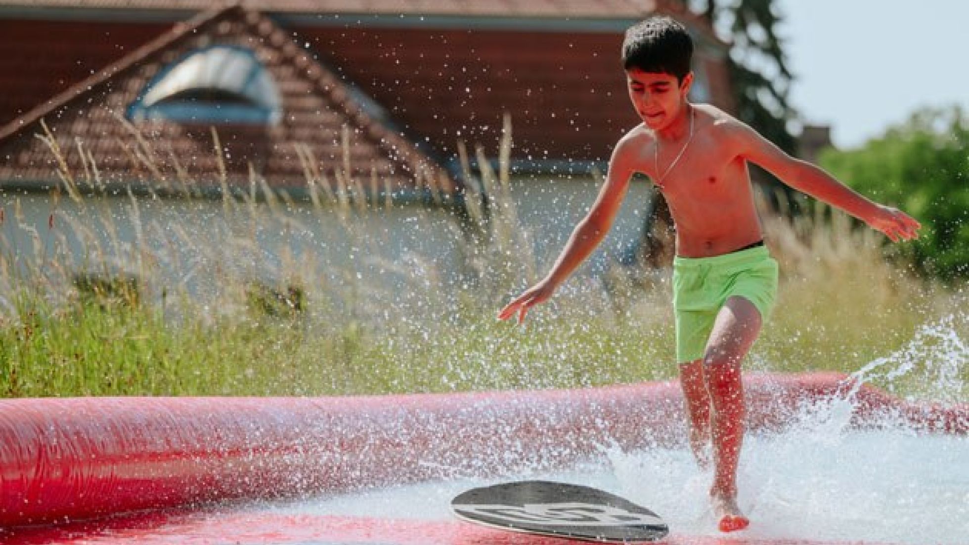 Auf einer Wiese ist eine rote Kunststoffplane ausgelegt und mit etwas Wasser befüllt. Ein Bub läuft durch das Wasser einem schwarzen Schwimmbrett hinterher.