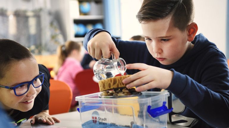 Ein Bub schüttet bei einem Experiment Wasser aus einem kleinen Krug auf einen Schwamm. Ein zweiter Bub sieht begeistert dabei zu.