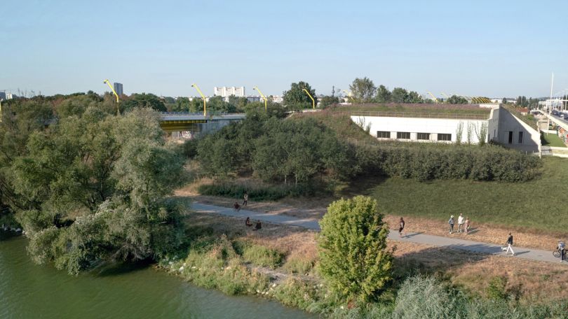 Zwischen 2 Brücken auf der Donauinsel steht ein niedriges weißes Gebäude mit begrüntem Dach. Davor wachsen Sträucher. Am Gehweg am Ufer spazieren Menschen.