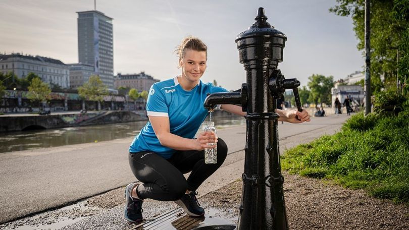 copyright: Karo Pernegger Eine Frau in Sportkleidung füllt ihre Wasserflasche bei einem Trinkwasserbrunnen auf.