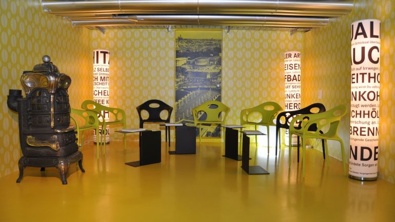 Gelb-grüner Ausstellungsraum mit Stühlen und einem Ofen