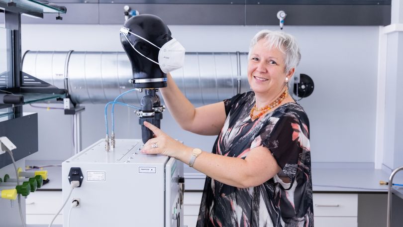 Mitarbeitern steht vor Maskenprüfgerät mit applizierter Atemschutzmaske und blickt lächelnd in die Kamera