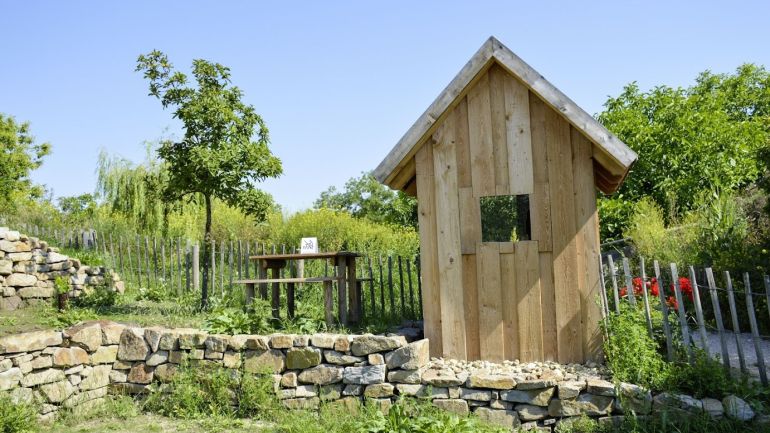 Kleine Holzhütte, daneben ein Tisch und Bänke, davor eine Steinmauer, dahinter ein Zaun, Bäume und Sträucher.