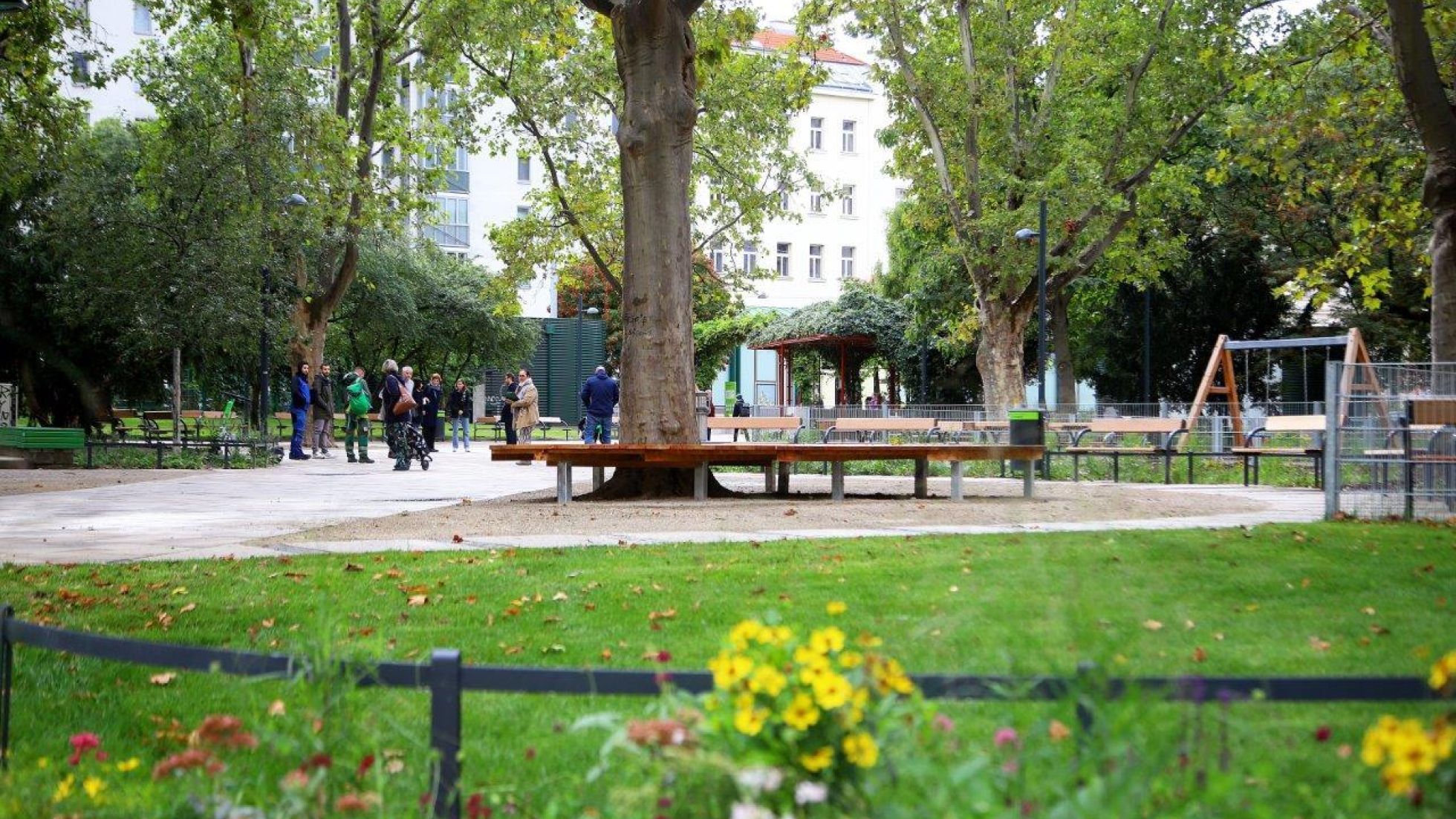 Zentraler Bereich in einem Park mit Sitzgelegenheiten und Spielplatz, im Vordergrund eine Wiese und Blumen