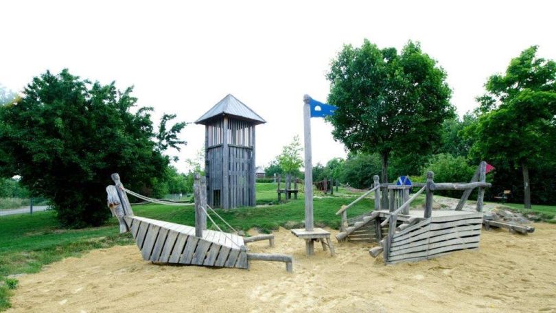 Sandkiste in Form eines Piratenschiffs aus Holz, rund herum Bäume und Wiese