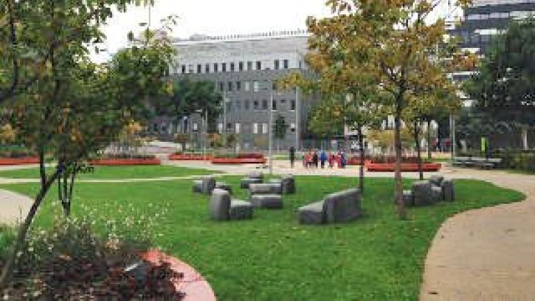 Wege führen durch Wiesen mit Sitzgelegenheiten, Bäume, Häuser im Hintergrund