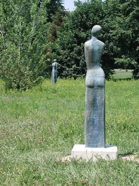 2 Frauenskulpturen in einer  Wiese mit hohem Gras, dahinter sind Bäume