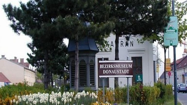 copyright: MA 42 Im Vordergrund ist ein buntes Frühlingsbeet mit einem Schild des Bezirksmuseums, dahinter ein kleines Haus