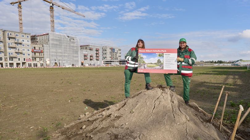 Eine Mitarbeiterin und ein Mitarbeiter der Stadtgärten halten eine Tafel zum neuen Albert-Schultz-Park und stehen auf einem Erdhaufen auf der Baustelle.