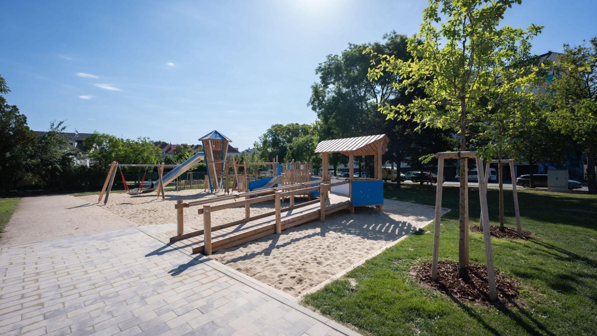 Spielplatz mit Sand, davor helle Pflasterung, daneben Wiese, Bäume und Hecken