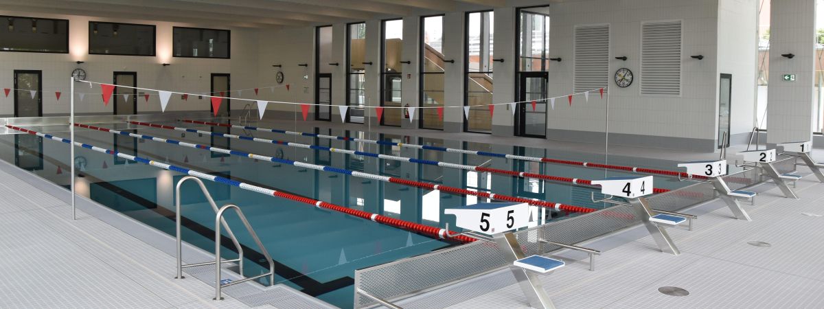 Ein Schwimmbecken mit Schwimmbahnen und Startblöcken. Über dem Wasser hängen rote und weiße Fähnchen.