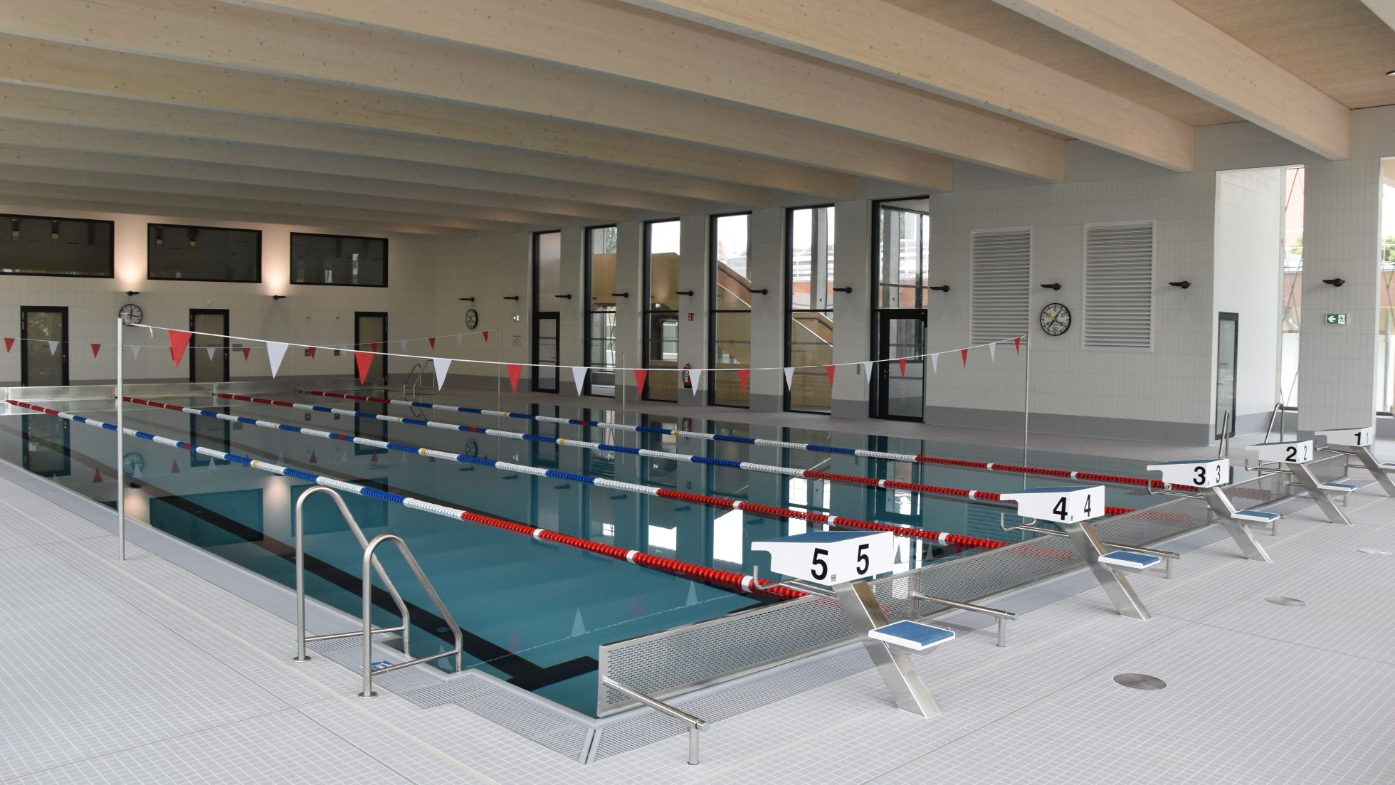 Ein Schwimmbecken mit Schwimmbahnen und Startblöcken. Über dem Wasser hängen rote und weiße Fähnchen.