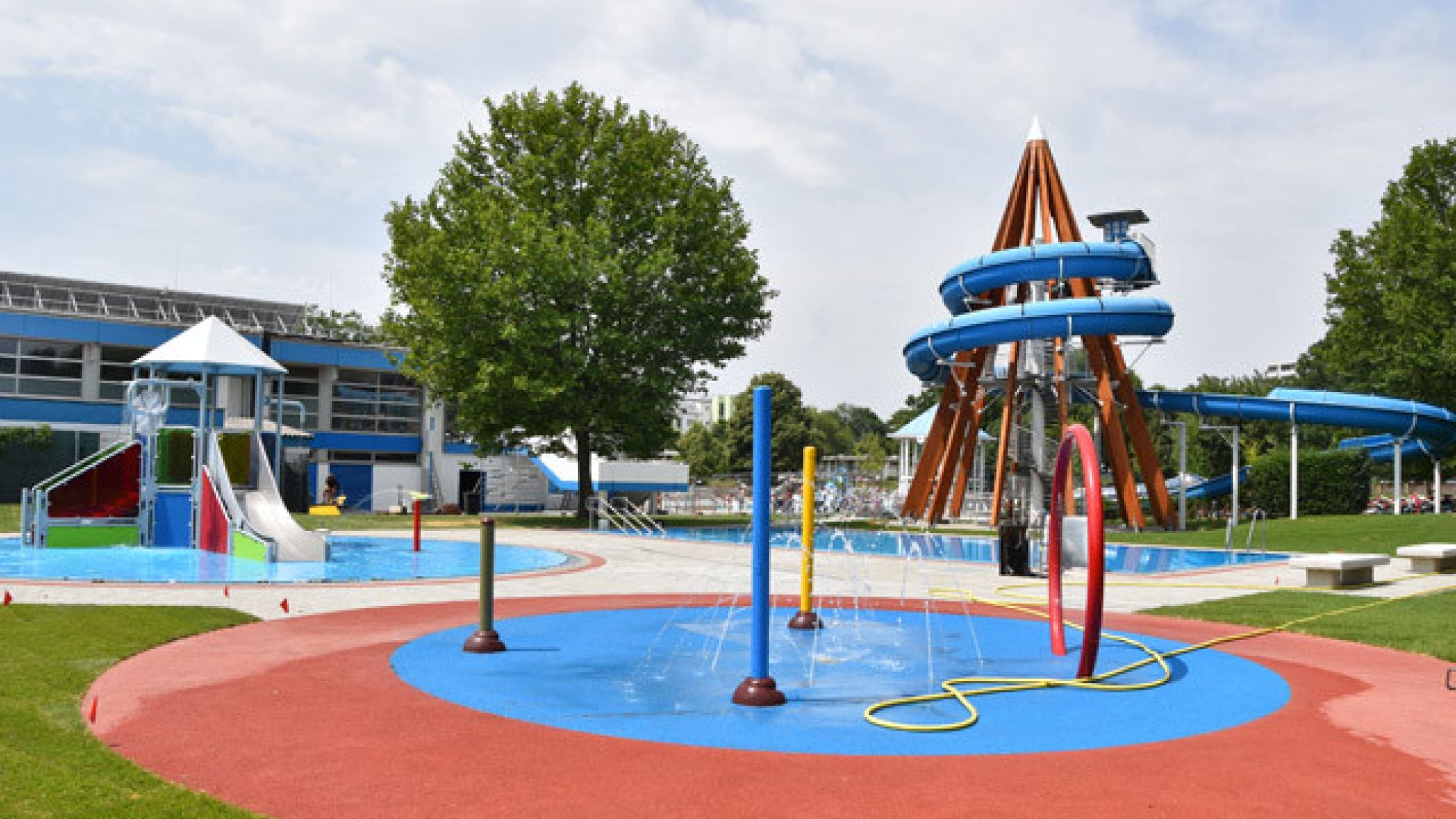 Wasserspielplatz mit Wasserfontänen, im Hintergrund ein hoher Turm mit Rutsche