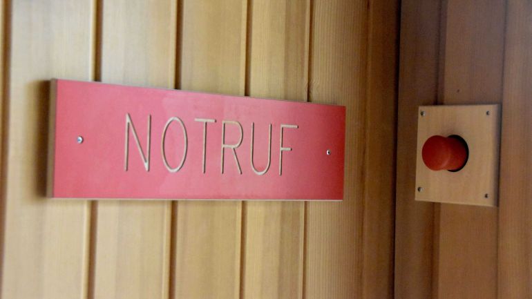 Roter Notfallknopf an der Wand. Daneben steht ihn Großbuchstaben "Notfallknopf".