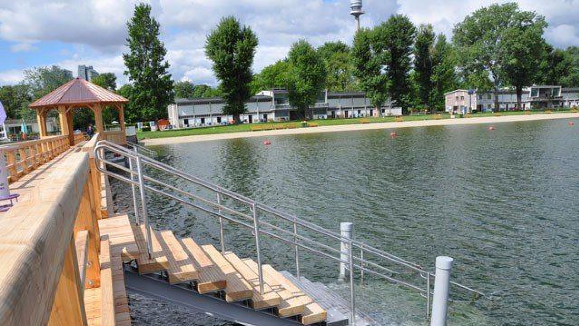 Vom Steg führen Stufen ins Wasser, im Hintergrund ist die Liegewiese am Ufer der Alten Donau zu sehen.