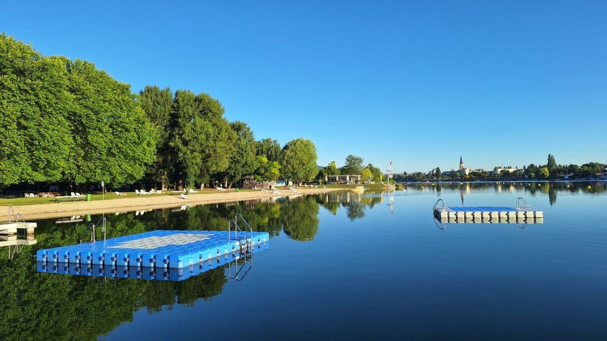 Eine blaue und eine weiße Badeinsel treiben im ruhigen Wasser. Am Ufer ist ein Sandstrand, dahinter große Bäume.