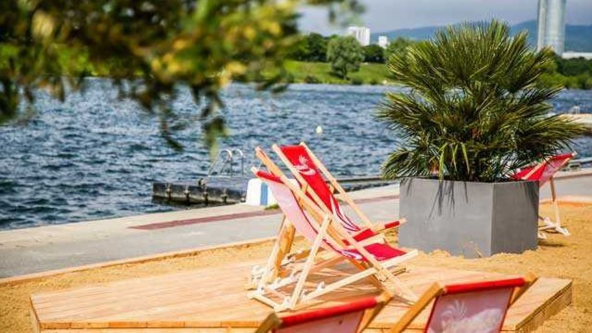 Am künstlichen Sandstrand stehen mehrere rote Liegestühle und eine Palme im Kübel.