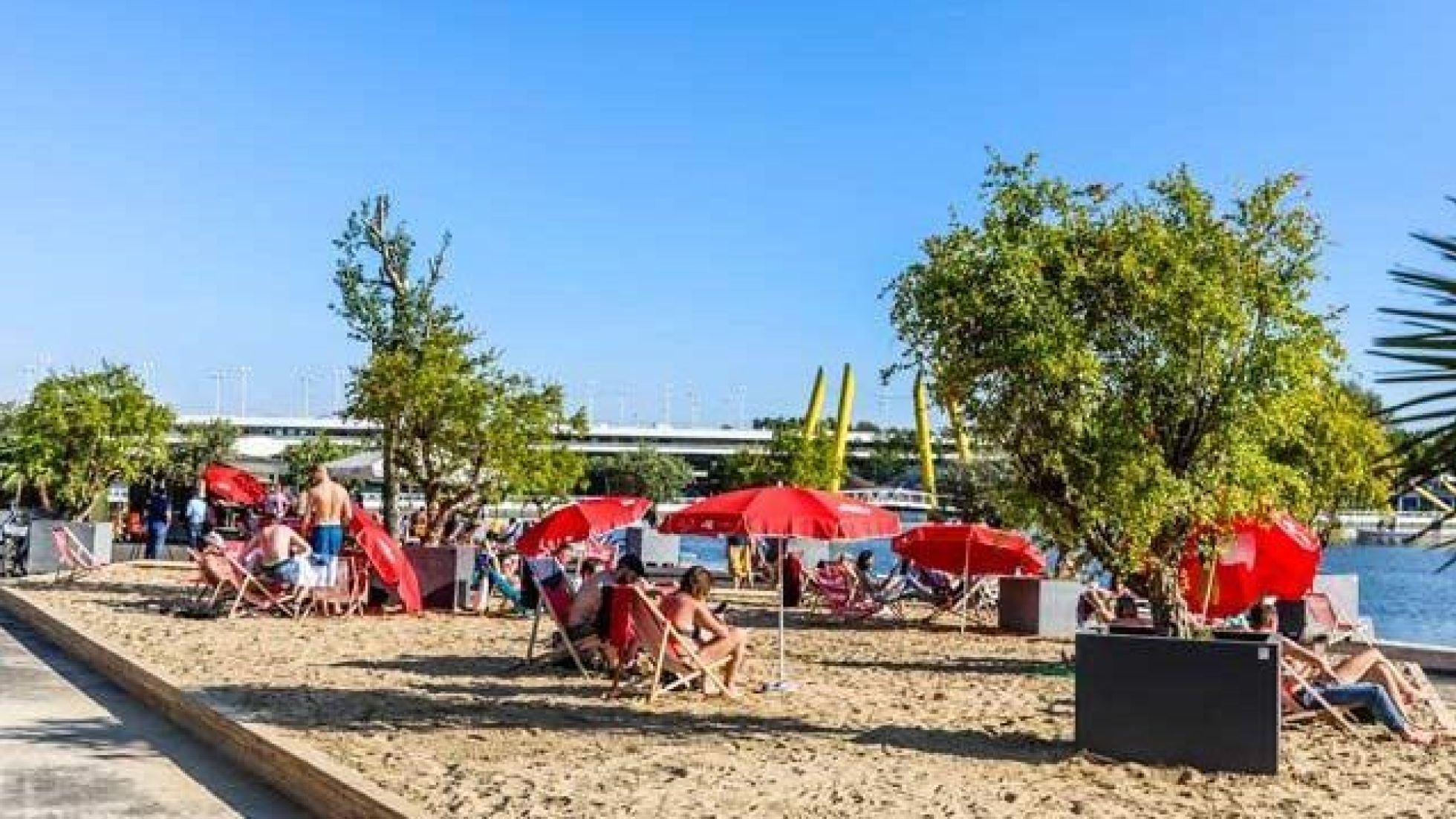 Menschen sitzen auf roten Liegestühlen unter roten Sonnenschirmen am künstlich angelegten Sandstrand.