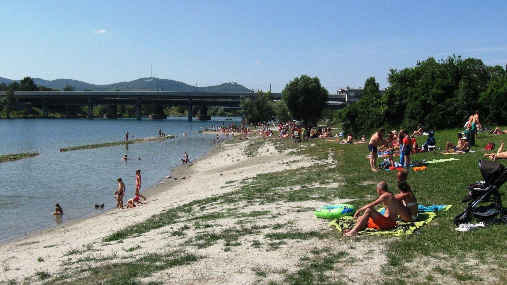 copyright: Wiener Gewässer (MA 45) Auf einer weitläufigen Liegewiese am Ufer der Neuen Donau sitzen viele Menschen in Badebekleidung auf Handtüchern. Einige Menschen gehen ins Wasser.