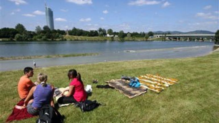 Drei Personen sitzen auf einer frisch gemähten Wiese, im Hintergrund Wasser, Bäume, eine Brücke und ein hohes Gebäude;