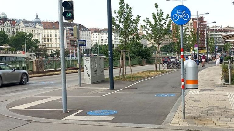 Radweg an einer Kreuzung. Der Radweg ist mit Verkehrszeichen und Piktogrammen am Boden gekennzeichnet und hat eine Fahrradampel, die grün anzeigt.