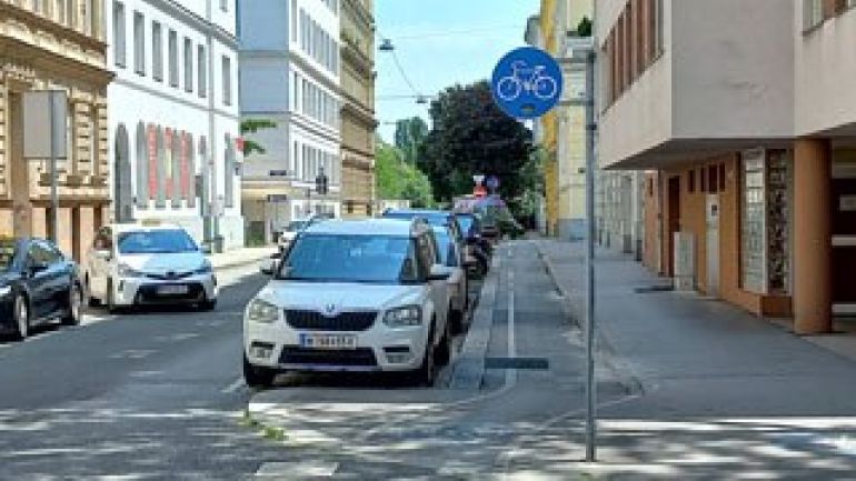 Ein Radweg verläuft zwischen Gehsteig und Parkspur. Der Radweg ist mit einem Verkehrszeichen gekennzeichnet, auf dem ein weißes Rad auf blauem Hintergrund zu sehen ist.