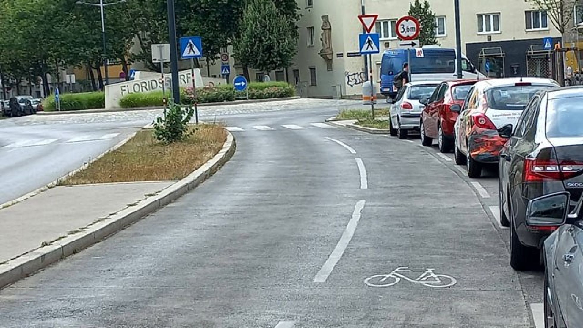 copyright: MA 46 Auf einer Straße ist ein Mehrzweckstreifen auf der Fahrbahn eingezeichnet. Rechts davon parken Autos. Die Straße mündet in einen Kreisverkehr.