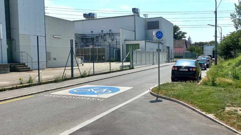 Auf der Fahrbahn ist ein großes Fahrradsymbol aufgezeichnet. Ein Verkehrsschild weißt auf die Fahrradstraße hin. Auf der rechten Seite parken Autos neben einer Wiese. Auf der linken Seite befindet sich ein Gehsteig und dahinter ein Industrie-Gelände.