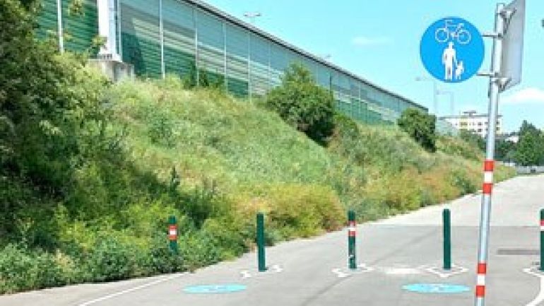 Der Geh- und Radweg ist mit einem Verkehrsschild und Piktogrammen am Boden gekennzeichnet. Boller verhindern die Einfahrt von Fahrzeugen. Links vom Weg ist eine Böschung.