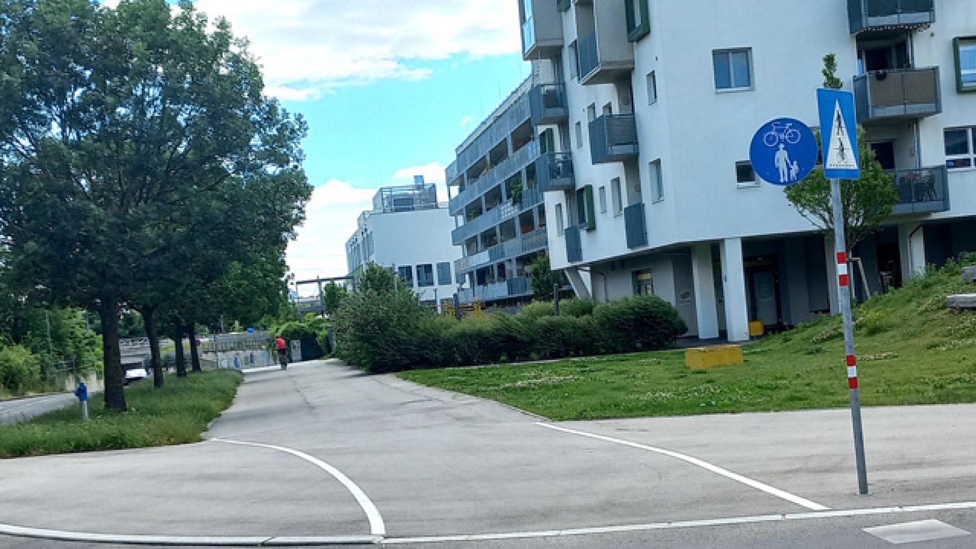 Zwischen einem Grünstreifen und einer Wohnhausanlage verläuft ein Geh- und Radweg. Der Weg ist mit einem Verkehrsschild gekennzeichnet.