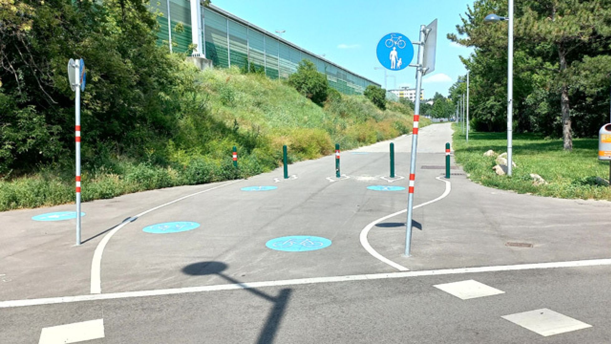 Am Boden sind Markierungen für einen Geh- und Radweg sowie eine Radfahrüberfahrt. Der Geh- und Radweg ist zusätzlich mit einem Verkehrszeichen gekennzeichnet.