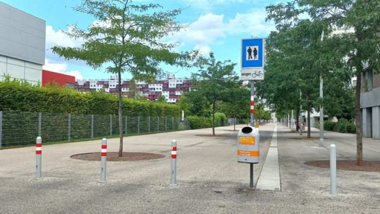 Ein Schild "Fußgängerzone ausgenommen Fahrräder" steht an einer Straße. Absperrpfosten blockieren die Zufahrt für große Fahrzeuge.