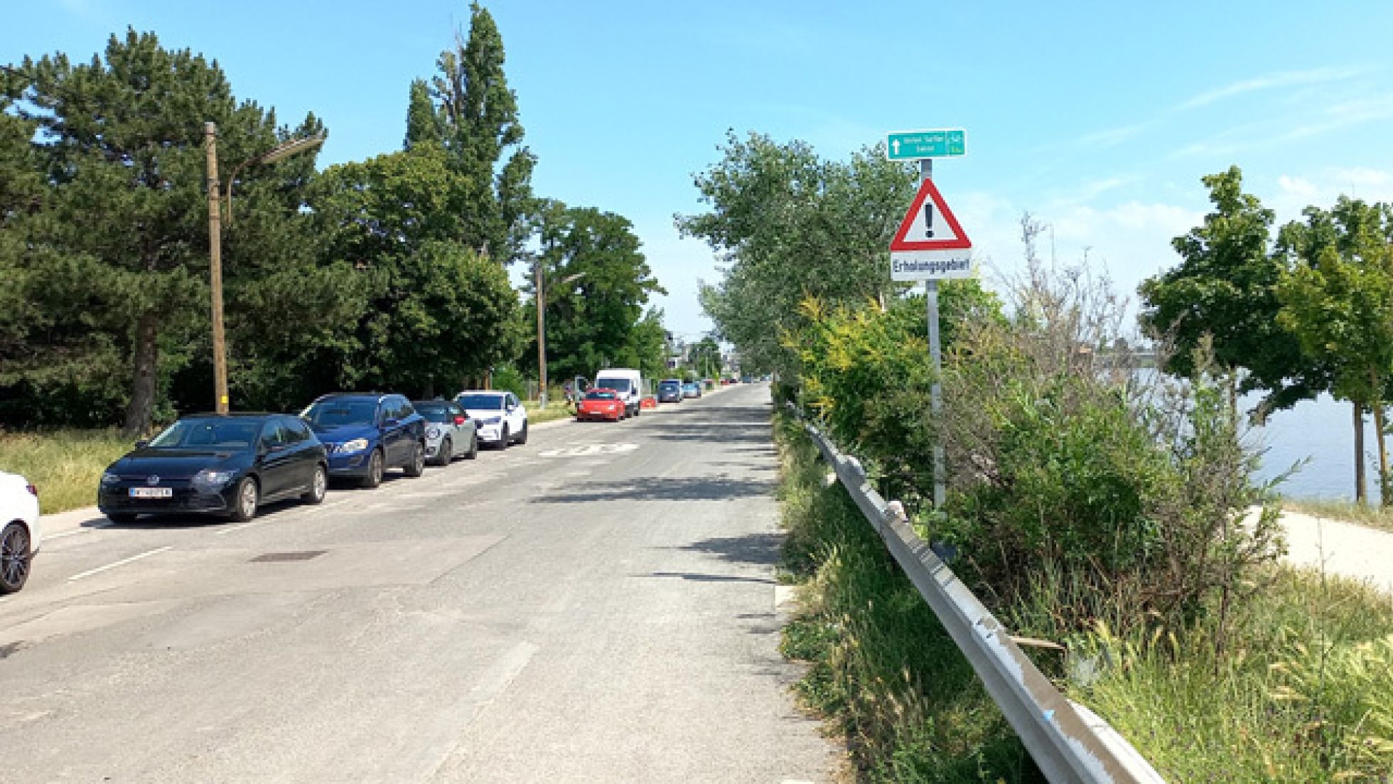 Rechts neben der Fahrbahn ist ein Hinweisschild für einen Radweg, darunter "Achtung" mit Zusatz "Erholungsgebiet". Auf einer Straßenseite parken Autos.