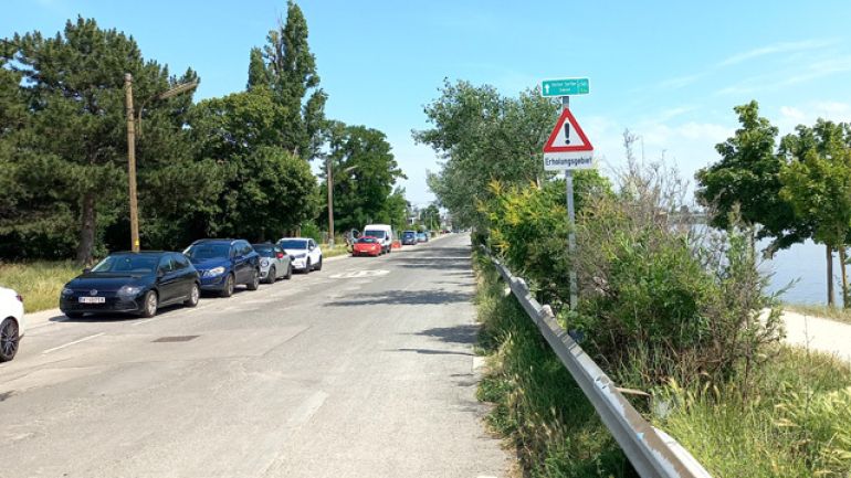 copyright: MA 46 Rechts neben der Fahrbahn ist ein Hinweisschild für einen Radweg, darunter "Achtung" mit Zusatz "Erholungsgebiet". Auf einer Straßenseite parken Autos.